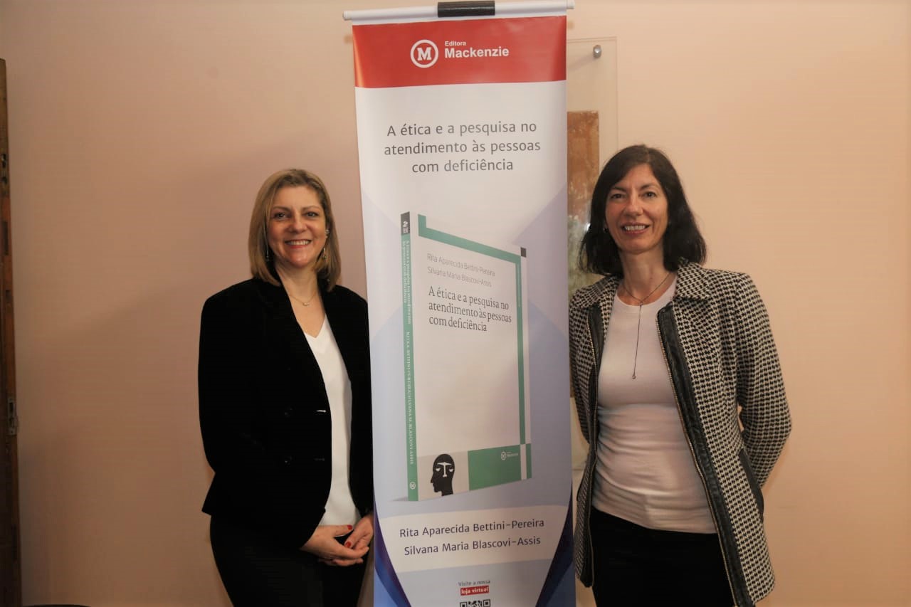 Autoras de pé ao lado do banner de apresentação do livro