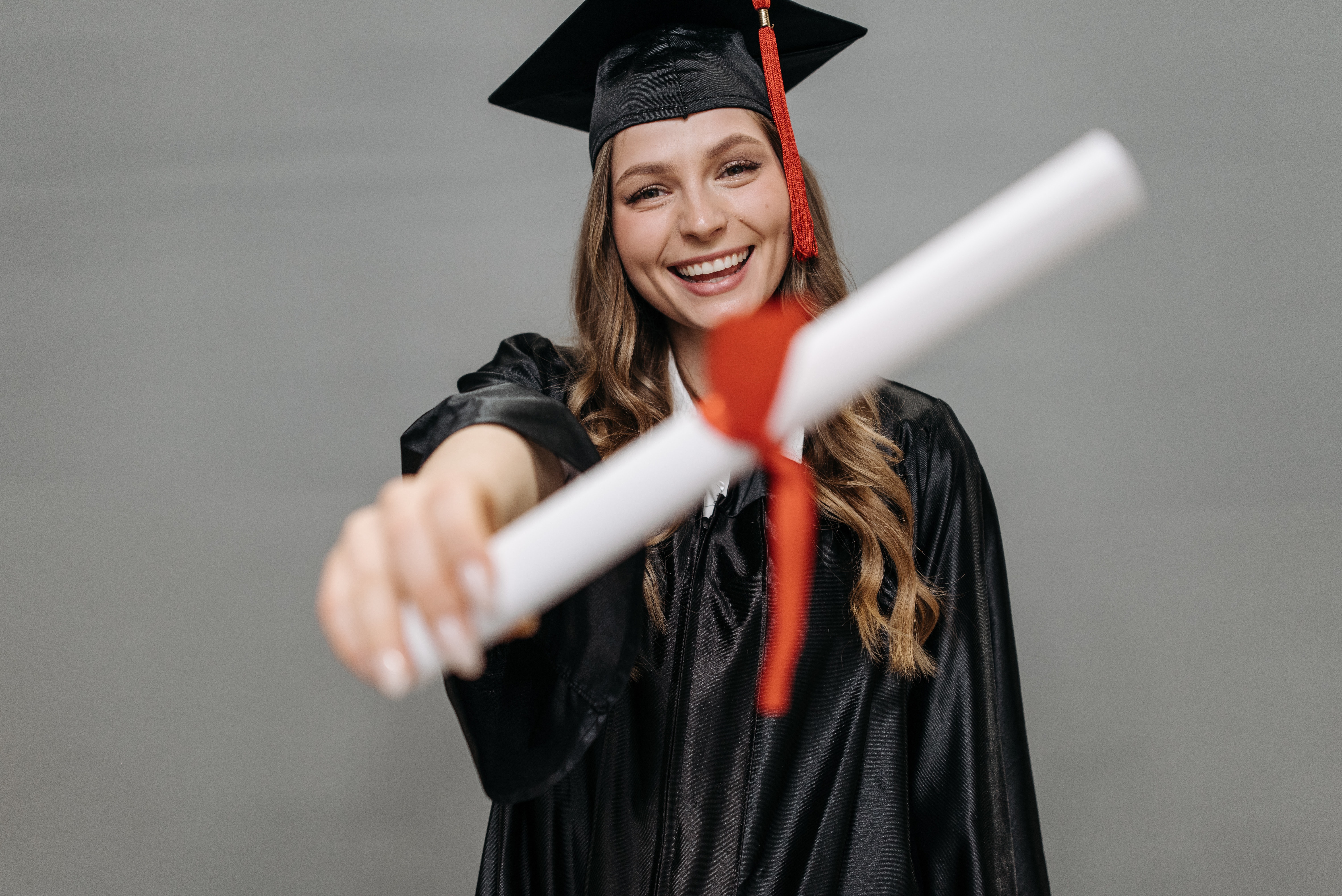 Na foto, uma menina mostra seu diploma para a câmera. Ela está de beca, com capelo e está sorrindo.
