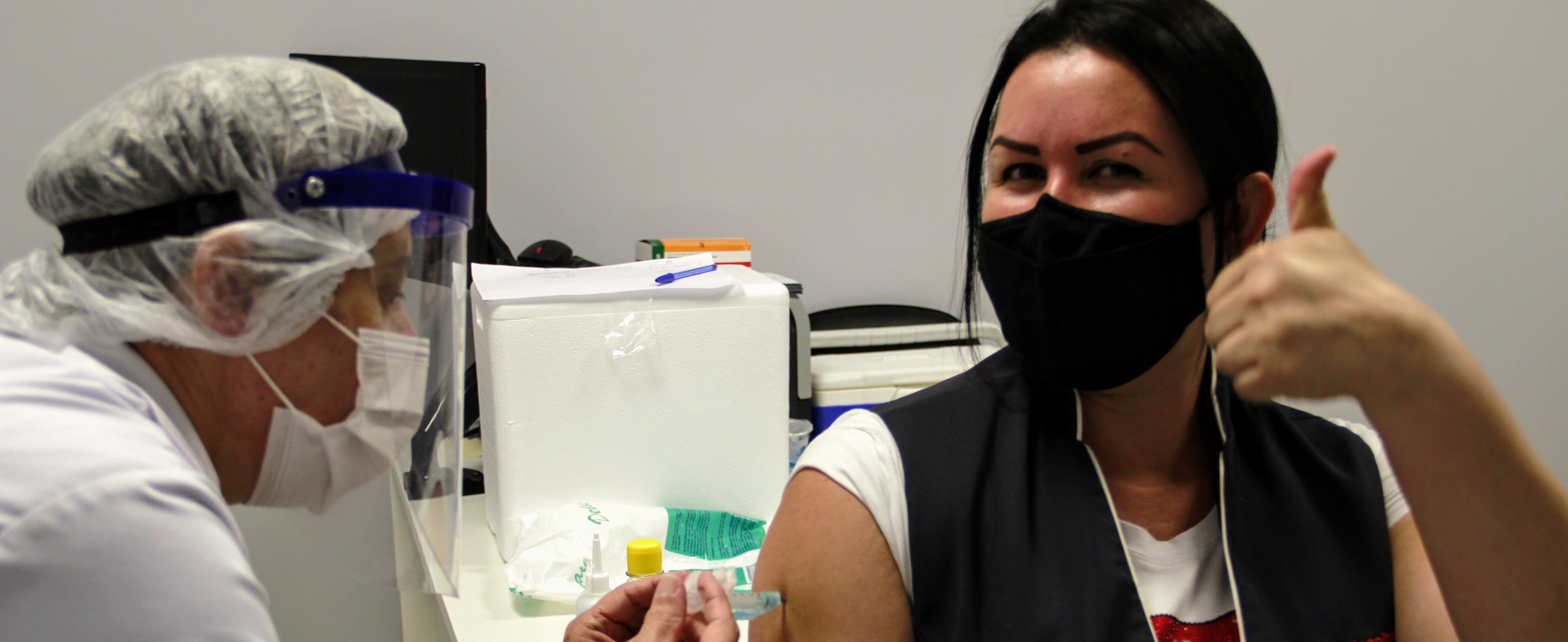 mulher morena, de máscara e com colete e crachá do MAckenzie, recebe injeção de outra mulher paramentada com máscara, jaleco e touca.