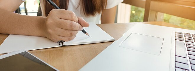 uma menina está escrevendo em um caderno, seu rosto não aparece. em sua frente tem um notebook