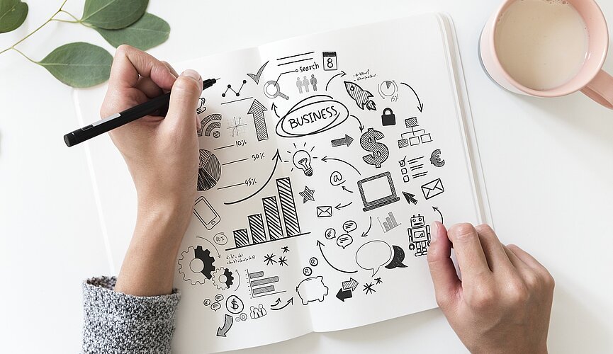 uma mão com um lápis sobre uma mesa. na folha de papel tem muitos desenhos referentes à economia