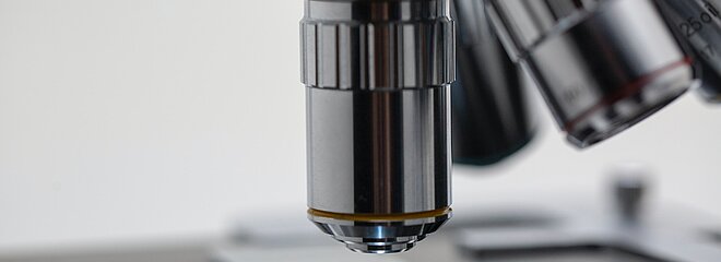 na imagem, uma foto bem próxima de uma das lentes de um microscópio nos tons cromado e prata analisando uma lâmina de vidro