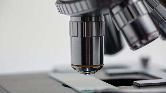 na imagem, uma foto bem próxima de uma das lentes de um microscópio nos tons cromado e prata analisando uma lâmina de vidro