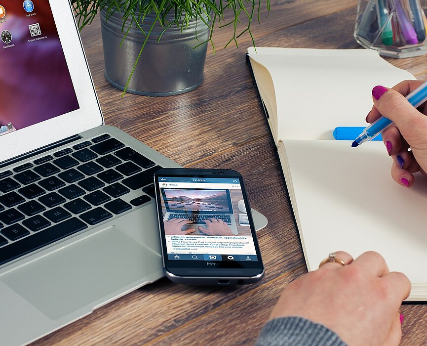 Mulher segurando um livrinho de notas, ao lado de um notebook e um celular