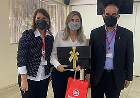 Duas mulheres brancas e um homem estão de pé posando para a foto. Os três estão vestidos de maneira social e usando máscaras