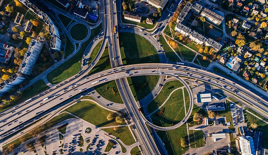 Foto aérea mostra estradas e diversos carros. Tem um pouco de verde e também algumas casas prédios, mas o foco são as vias com veículos.