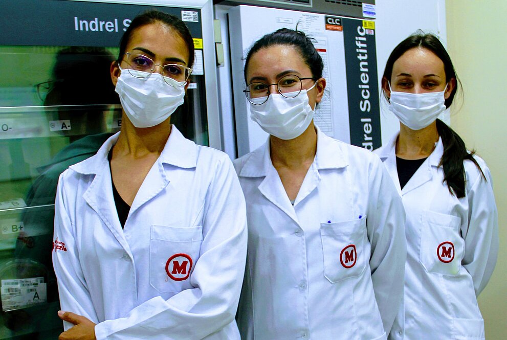 três mulheres de pé, em jalecos brancos, usando máscaras, posam para foto. Ao fundo, há refrigeradores de armazenamento de sangue