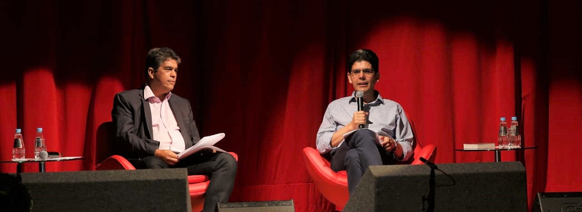 Vice-reitor e reverendo sentados com microfones em mãos durante apresentação, ao fundo, uma cortina vermelha.