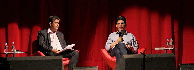 Vice-reitor e reverendo sentados com microfones em mãos durante apresentação, ao fundo, uma cortina vermelha.
