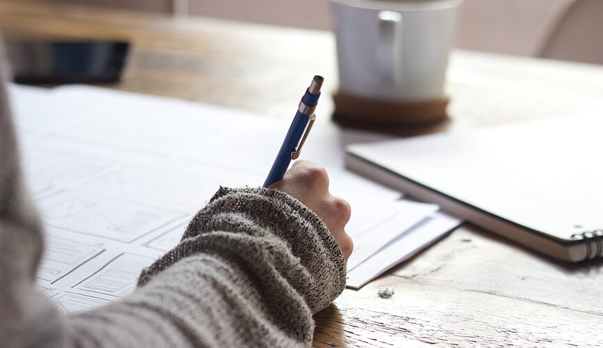 mão de garota segurando lápis e escrevendo em um caderno sobre uma mesa com uma xícara ao lado