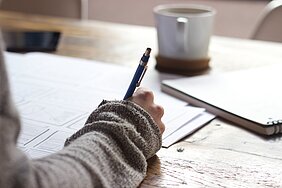 mão de garota segurando lápis e escrevendo em um caderno sobre uma mesa com uma xícara ao lado
