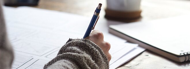 mão de garota segurando lápis e escrevendo em um caderno sobre uma mesa com uma xícara ao lado