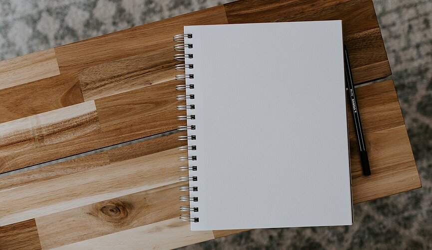A foto mostra um caderno em cima de uma mesinha de madeira. Ao lado tem uma caneta. 