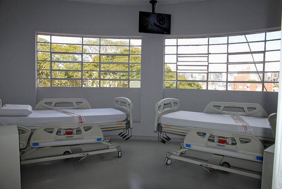 quarto hospitalar com camas e televisor, ao fundo, uma janela grande