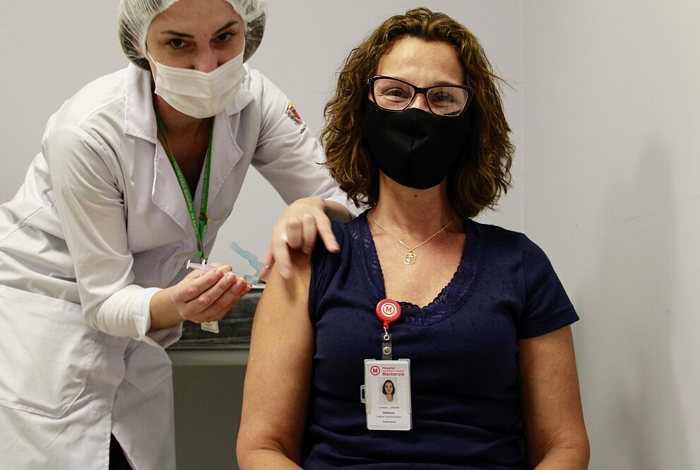 Mulher branca, sentada, de blusa preta, recebe vacina no braço de mulher de pé paramentada com jaleco, touca e luvas. Ambas estão de máscara.
