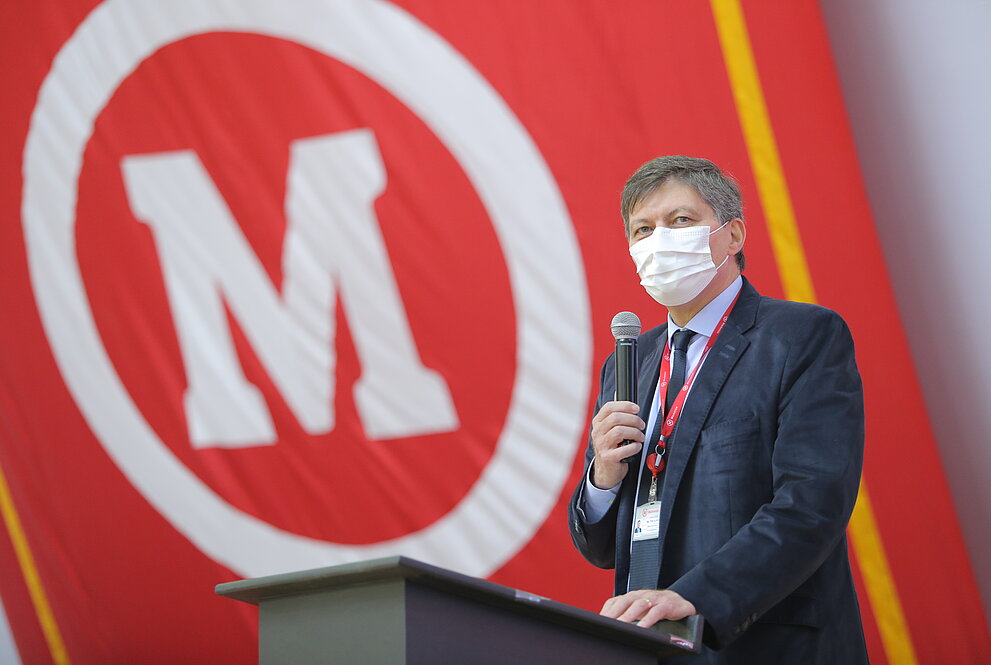 Homem de terno falando ao microfone com bandeira do mackenzie ao fundo