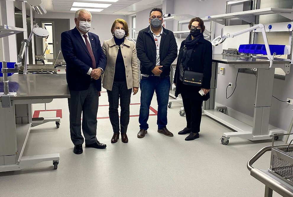 dois homens e duas mulheres de máscara estão de pé em uma sala de esterilização de materiais onde se pode ver objetos hospitalares novos