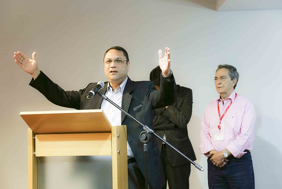 reverendo Carlos ora de mãos estendidas em frente ao púlpito. duas pessoas estão atrás dele