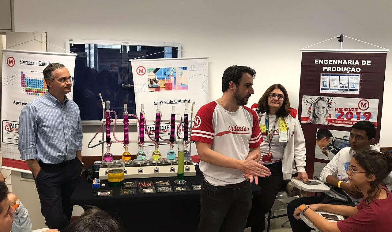 Pessoas de pé explicam sobre o curso aos visitantes sentados. Ao fundo há materiais de laboratório químico com diversas cores