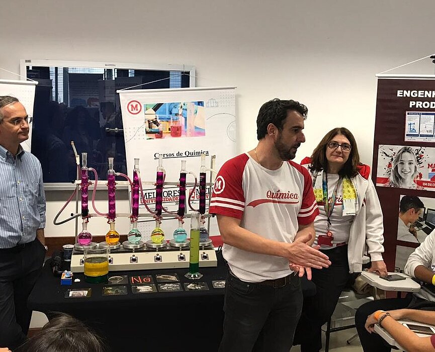 Pessoas de pé explicam sobre o curso aos visitantes sentados. Ao fundo há materiais de laboratório químico com diversas cores