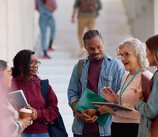 A imagem contem um grupo de jovens estudantes escutando sua professora em um local aberto.