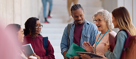A imagem contem um grupo de jovens estudantes escutando sua professora em um local aberto.