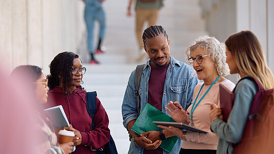 A imagem contem um grupo de jovens estudantes escutando sua professora em um local aberto.