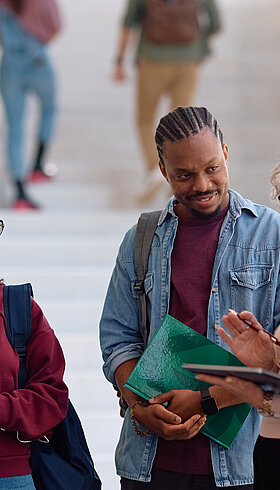 A imagem contem um grupo de jovens estudantes escutando sua professora em um local aberto.