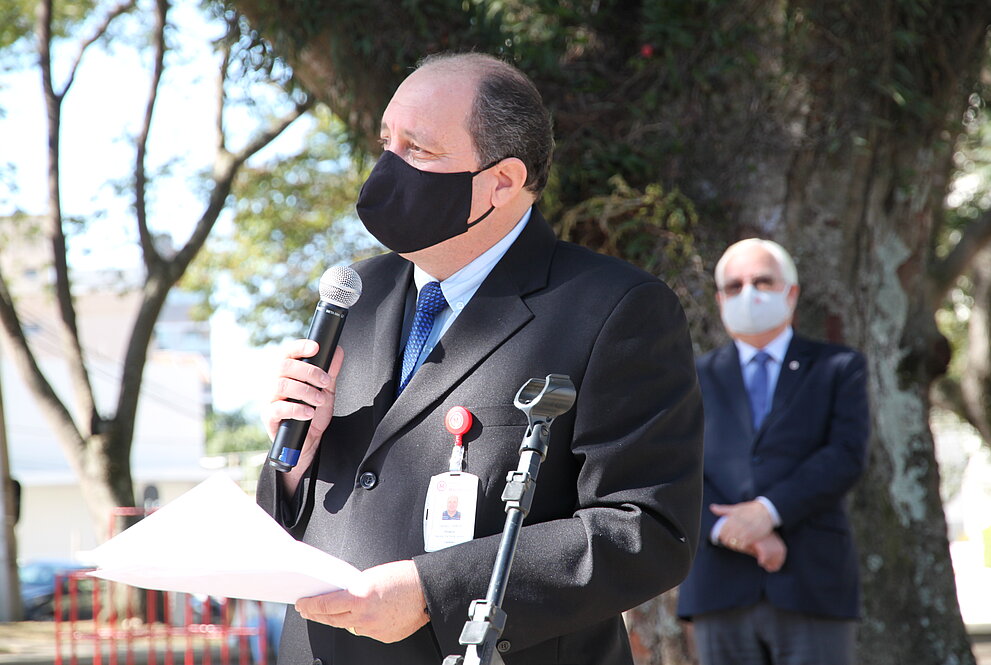 Homem de máscara, terno e com microfone, discursa a céu aberto com árvores ao fundo