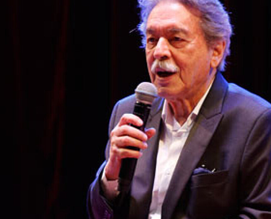 A foto mostra o arquiteto Paulo Mendes da Rocha em um evento do Mackenzie, no ano de 2017. Ele está com um blazer e segurando um microfone, sentado em uma poltrona vermelha. 