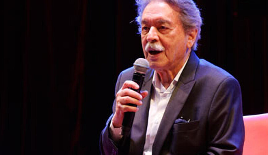 A foto mostra o arquiteto Paulo Mendes da Rocha em um evento do Mackenzie, no ano de 2017. Ele está com um blazer e segurando um microfone, sentado em uma poltrona vermelha.