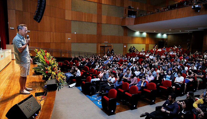 Thái à esquerda, sobre o palco, conversa com auditório cheio, à direita.