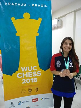 Julia Alboredo ao lado de banner da competição WUC Chess 2018