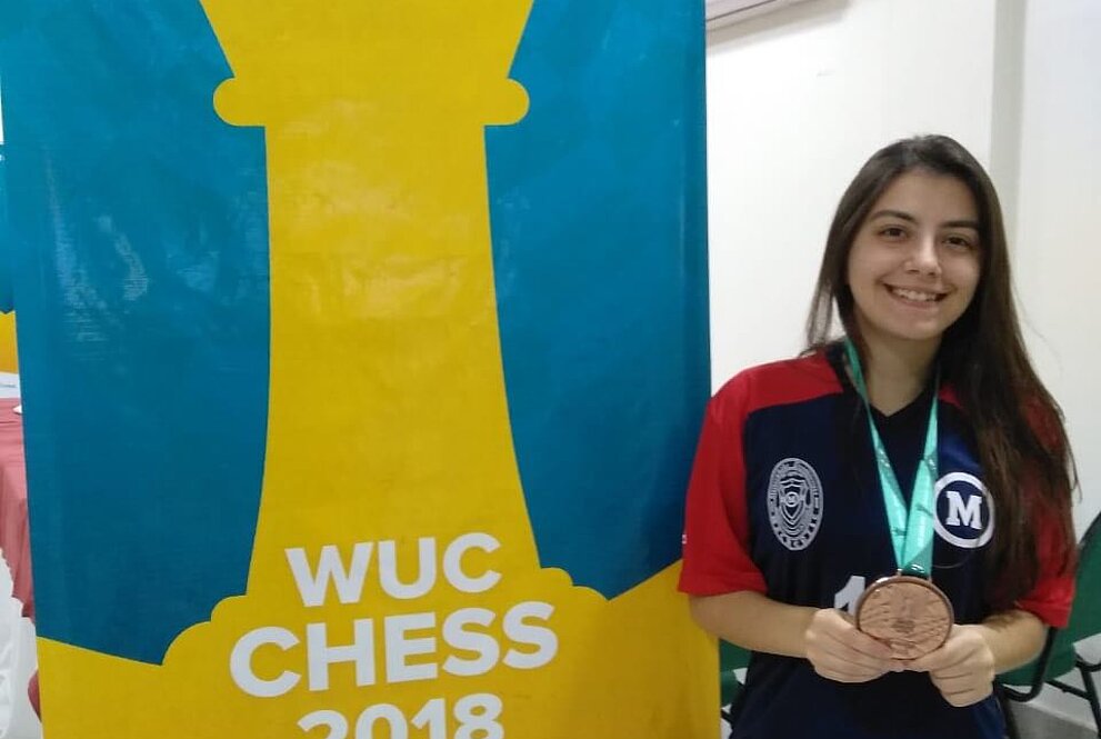 Julia Alboredo ao lado de banner da competição WUC Chess 2018