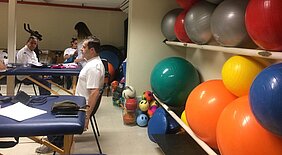 Do lado direito da imagem tem uma parede com bolas de ginástica de várias cores e do lado direito está um homem sentado fazendo exercícios ao mesmo tempo que outro homem também faz no fundo da imagem