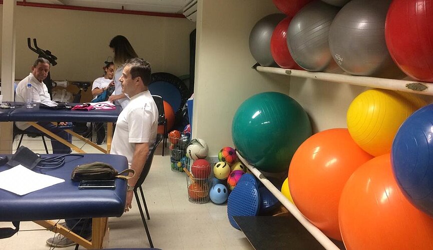 Do lado direito da imagem tem uma parede com bolas de ginástica de várias cores e do lado direito está um homem sentado fazendo exercícios ao mesmo tempo que outro homem também faz no fundo da imagem