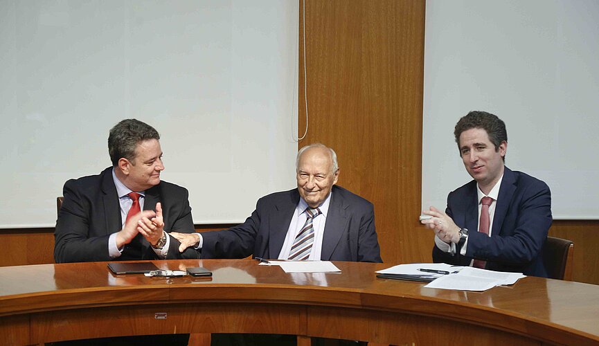 Três homens sentados ao redor de uma mesa redonda de madeira se cumprimentam.