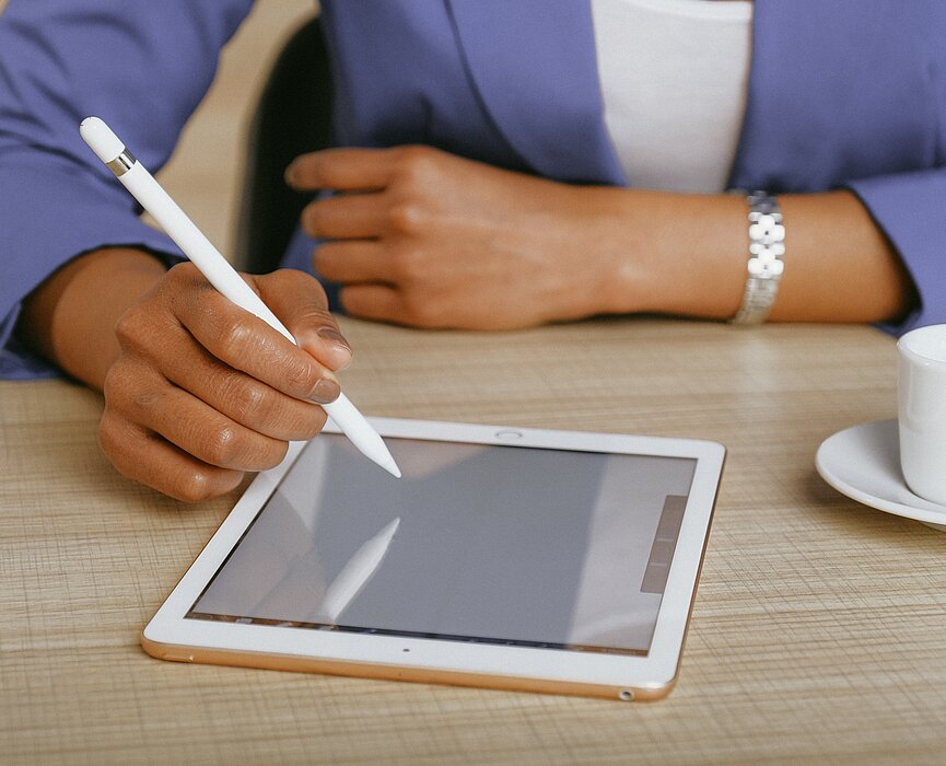 A foto mostra os braços de uma mulher apoiados sobre uma mesa. Ela está usando um tablet, com uma caneta. Uma xícara também está ao seu lado. Ela está usando um blazer, o que remete à negócios. 