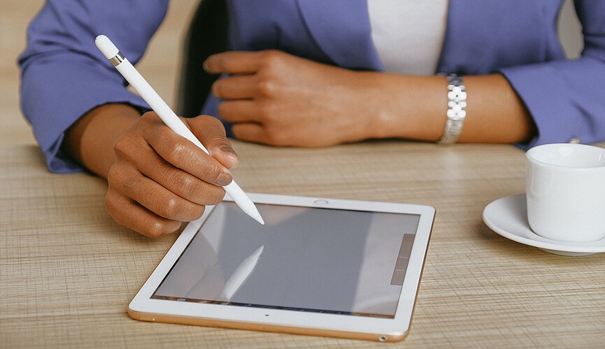 A foto mostra os braços de uma mulher apoiados sobre uma mesa. Ela está usando um tablet, com uma caneta. Uma xícara também está ao seu lado. Ela está usando um blazer, o que remete à negócios. 