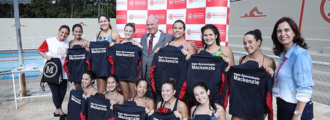 Atletas do nado sincronizado de Brasília enfileiradas em frente à piscina segurando seus agasalhos esportivos pretos do Mackenzie.