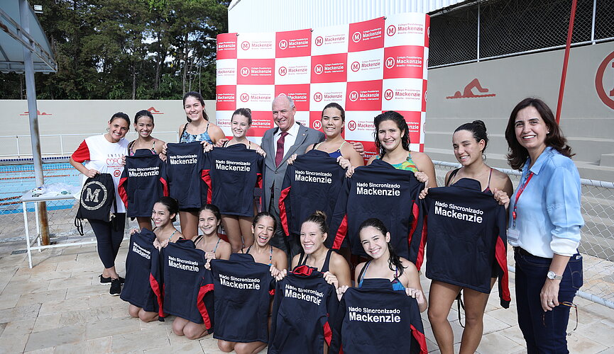 Atletas do nado sincronizado de Brasília enfileiradas em frente à piscina segurando seus agasalhos esportivos pretos do Mackenzie.