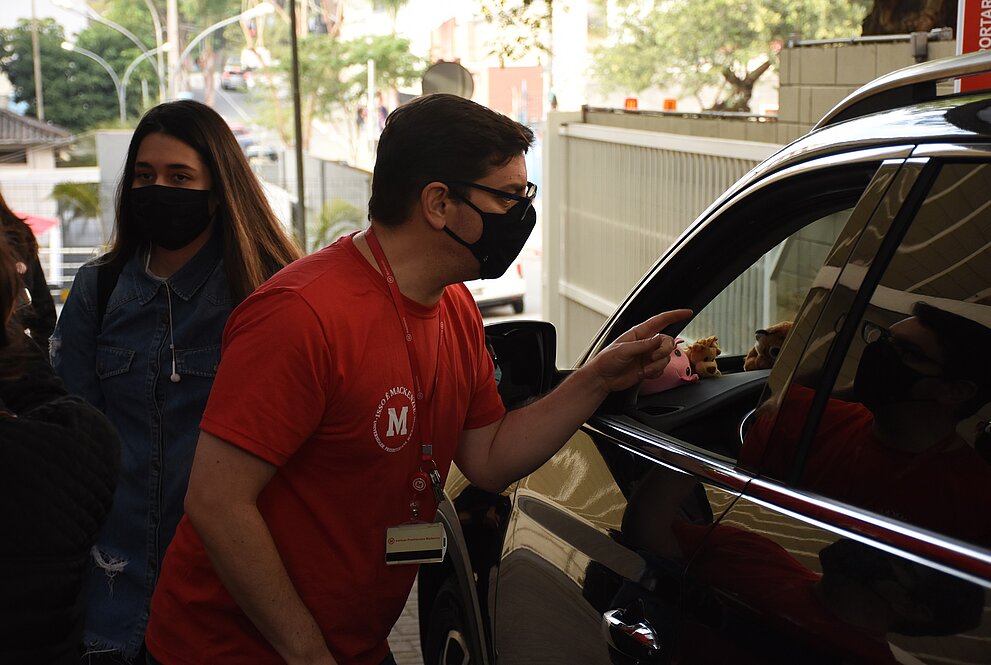 Homem de camiseta vermelha e máscara conversa com motorista de carro da cor preta
