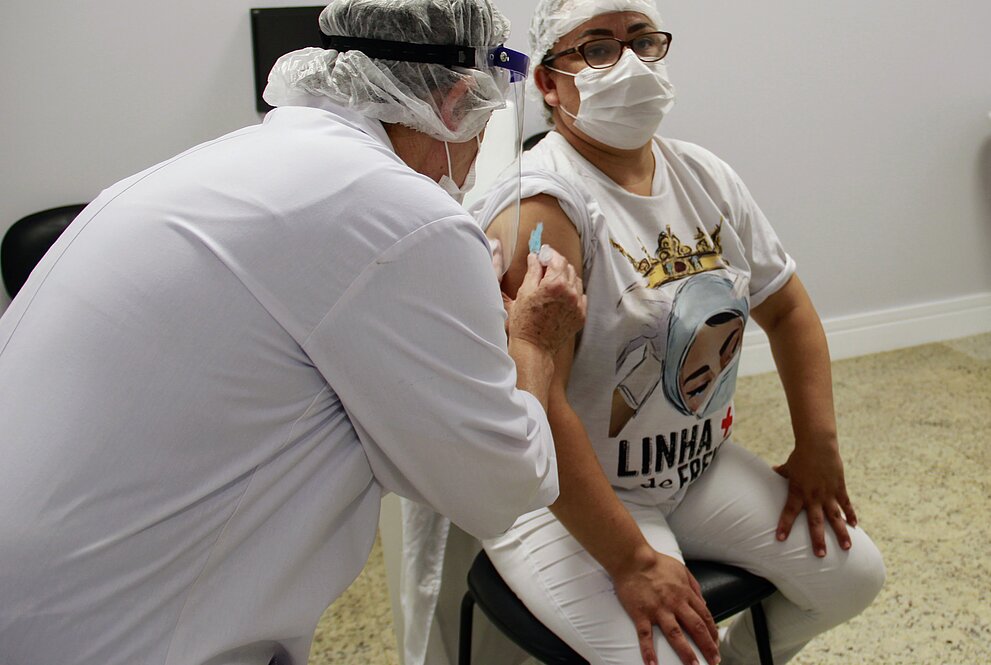 Profissional da saúde de pé, visto de costas, aplica vacina em colaboradora sentada. Ambos estão de máscara.