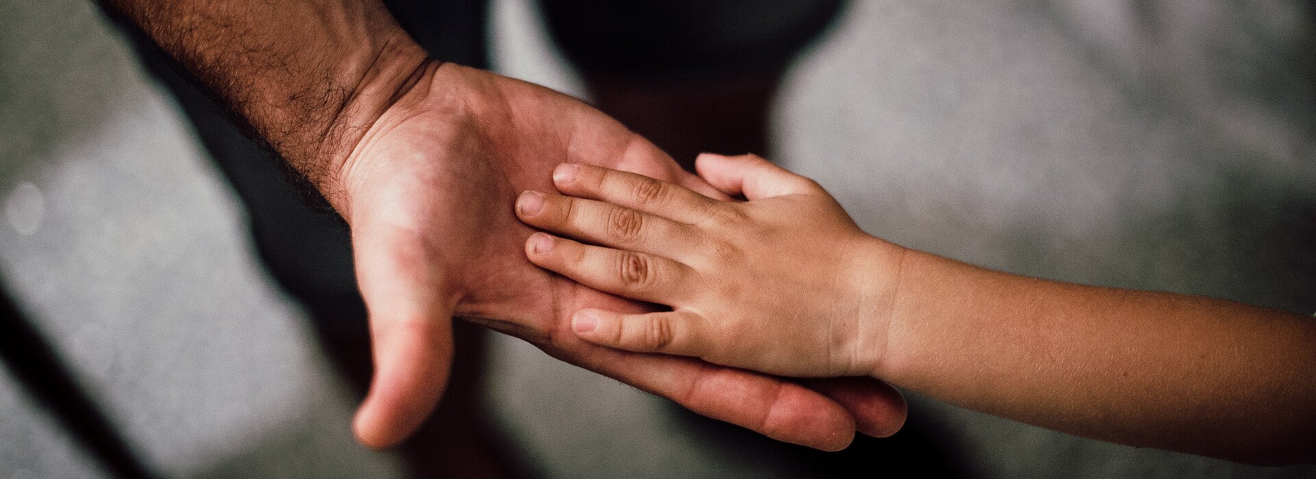 Um adulto estica a mão para uma criança. A foto tem foco na mão deles. 