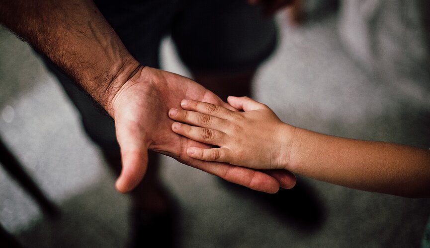 Um adulto estica a mão para uma criança. A foto tem foco na mão deles. 