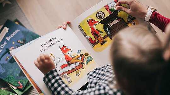A foto foi tirada de cima e mostra a cabeça de uma criança e suas mãos em cima de um livro, com ilustrações de uma raposa. A criança está acompanhando por um adulto, porque conseguimos ver a mão de uma mulher adulta, ajudando a criança na leitura.