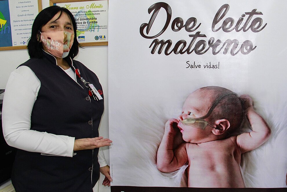 mulher branca, de blusa branca e colete escuro posa ao lado de um banner onde se vê um bebê recém-nascido e os dizeres "doe leite materno"