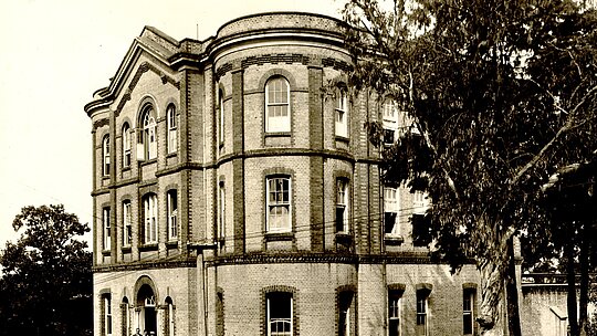 A foto é antiga e mostra o prédio Chamberlain, do campus Higienópolis. Do lado direito, na frente do prédio, tem uma árvore. Já do lado esquerdo, uma árvore está ao fundo da imagem. 