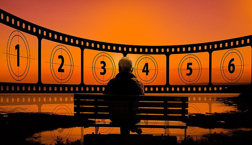 Idoso sentado em um banco de parque, em frente a um lago, com horizonte com vista de rolo de filmes numerado. Foto preta e laranja.