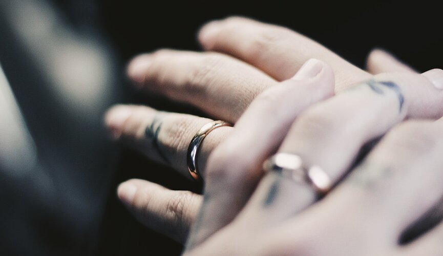 a foto mostra a mão de duas pessoas em close usando alianças de casamento e sobrepostas uma a outra